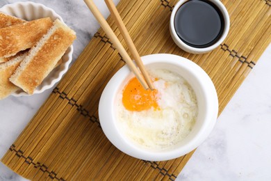 Photo of Half-boiled egg in bowl, soy sauce, chopsticks and toasted bread on white marble table, flat lay