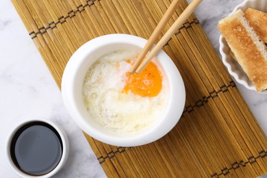 Photo of Half-boiled egg in bowl, soy sauce, chopsticks and toasted bread on white marble table, flat lay