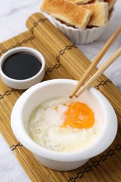 Photo of Half-boiled egg in bowl, soy sauce, chopsticks and toasted bread on white marble table, closeup