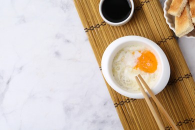 Photo of Half-boiled egg in bowl, soy sauce, chopsticks and toasted bread on white marble table, flat lay. Space for text