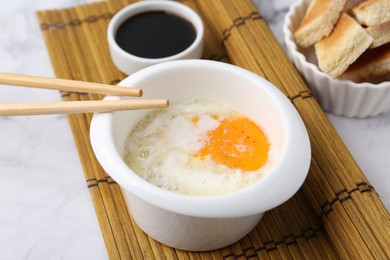 Photo of Half-boiled egg in bowl, soy sauce, chopsticks and toasted bread on white marble table, closeup