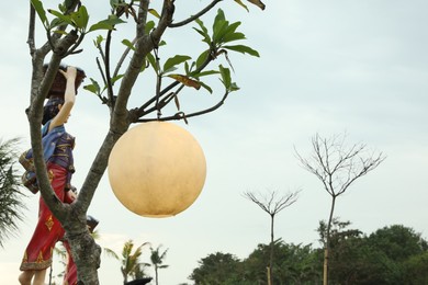 Photo of Bali, Indonesia - May 27, 2025: Beautiful lantern hanging on tree and traditional statue outdoors, low angle view