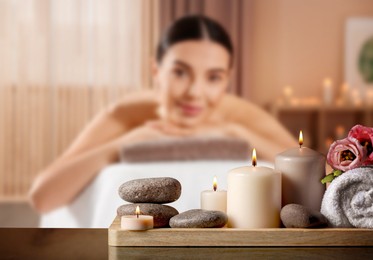 Image of Spa therapy. Woman lying on massage table and tray with hot stones and burning candles in salon, selective focus