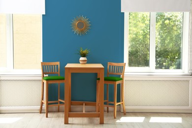 Image of Dining table and chairs near blue wall and windows in room. Interior design