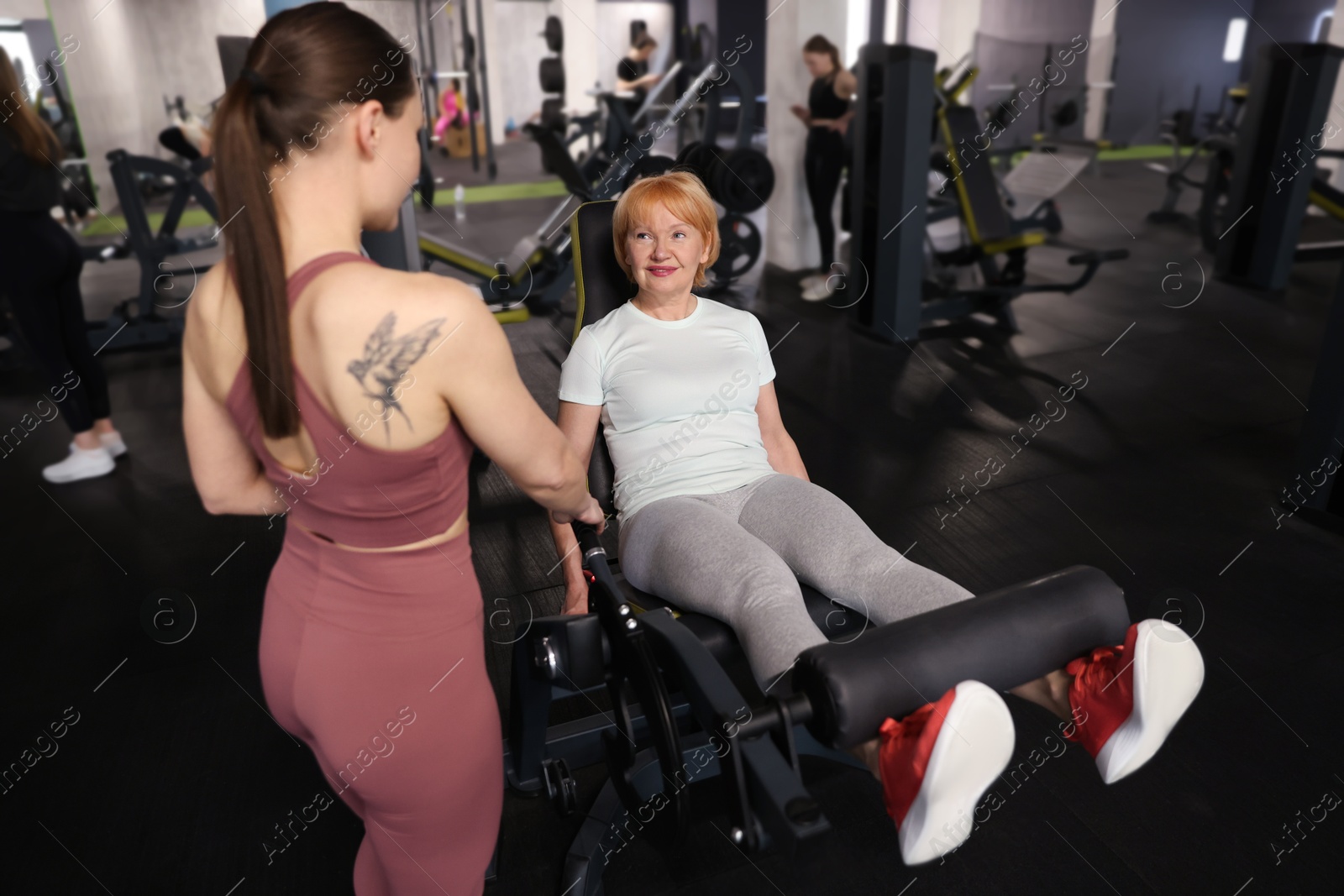 Senior woman training on leg extension machine supervised by professional trainer in gym Photo of Senior woman training on leg extension machine supervised by professional trainer in gym