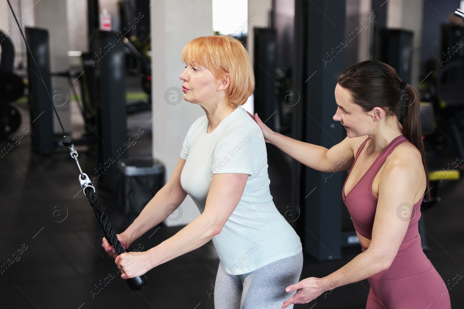 Senior woman training on pulley machine supervised by professional trainer in gym Photo of Senior woman training on pulley machine supervised by professional trainer in gym