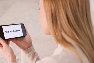 Photo of Cyber bullying. Upset teenage girl looking at smartphone with text You Loser at home, closeup