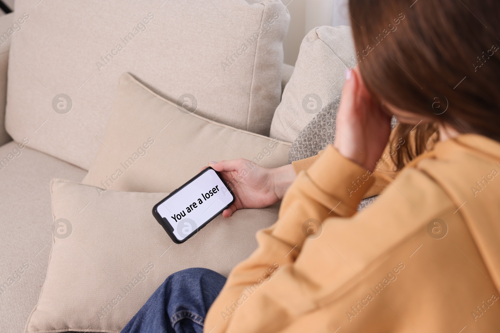 Cyber bullying. Upset teenage girl looking at smartphone with text You Loser on sofa at home, closeup Photo of Cyber bullying. Upset teenage girl looking at smartphone with text You Loser on sofa at home, closeup