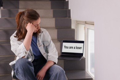 Photo of Cyber bullying. Upset teenage girl and laptop with text You Loser on stairs indoors