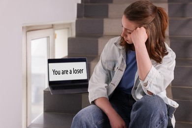 Photo of Cyber bullying. Upset teenage girl and laptop with text You Loser on stairs indoors