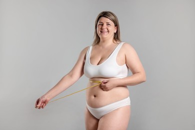 Photo of Weight loss. Plus size woman measuring waist with tape on gray background