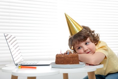 Photo of Birthday celebration. Little boy with cake having video call via laptop at table indoors. Space for text