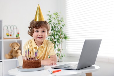 Photo of Birthday celebration. Little boy with cake having video call via laptop at table indoors. Space for text