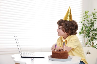 Photo of Birthday celebration. Little boy with cake having video call via laptop at table indoors. Space for text