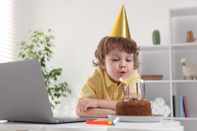 Photo of Birthday celebration. Little boy blowing candles on cake while having video call via laptop at table indoors