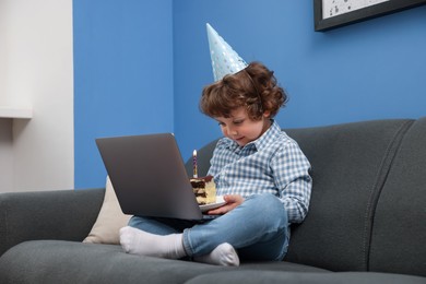 Photo of Birthday celebration. Little boy with piece of cake having video call via laptop on sofa at home