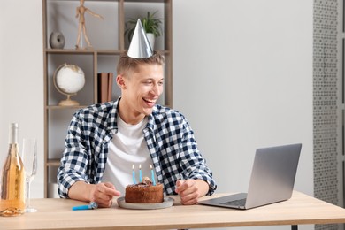 Photo of Birthday celebration. Man with cake and sparkling wine having video call via laptop at desk indoors