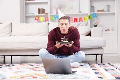 Photo of Birthday celebration. Man blowing candle on piece of cake while having video call via laptop at home