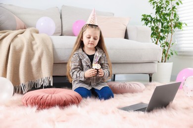 Photo of Birthday celebration. Little girl with cupcake having video call via laptop at home