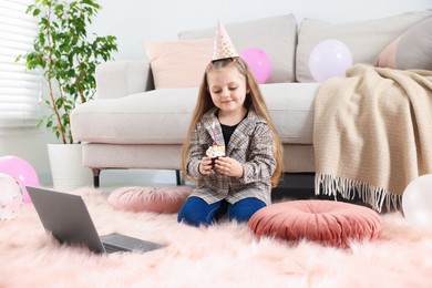 Photo of Birthday celebration. Little girl with cupcake having video call via laptop at home