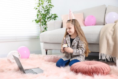 Photo of Birthday celebration. Little girl with cupcake having video call via laptop at home