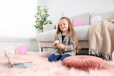 Photo of Birthday celebration. Little girl with cupcake having video call via laptop at home