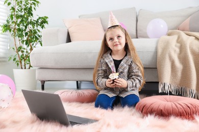 Photo of Birthday celebration. Little girl with cupcake having video call via laptop at home