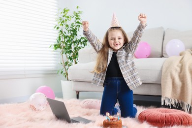 Photo of Birthday celebration. Little girl with cake having video call via laptop at home