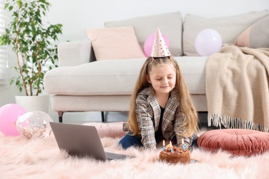 Photo of Birthday celebration. Little girl with cake having video call via laptop at home