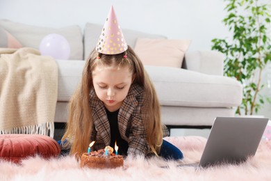 Photo of Birthday celebration. Little girl blowing candles on cake having video call via laptop at home