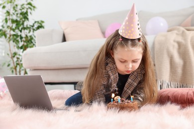 Photo of Birthday celebration. Little girl blowing candles on cake having video call via laptop at home