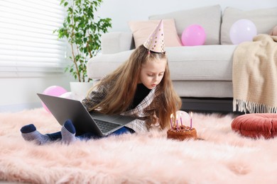 Photo of Birthday celebration. Little girl blowing candles on cake having video call via laptop at home