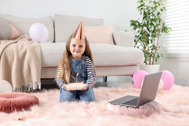 Photo of Birthday celebration. Little girl with piece of cake having video call via laptop at home