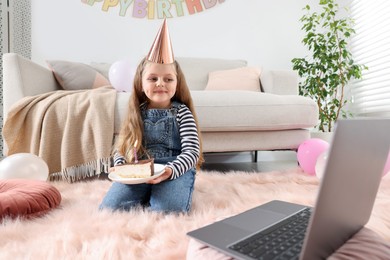 Photo of Birthday celebration. Little girl with piece of cake having video call via laptop at home