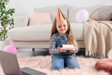 Photo of Birthday celebration. Little girl with piece of cake having video call via laptop at home
