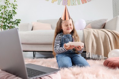Photo of Birthday celebration. Little girl with piece of cake having video call via laptop at home