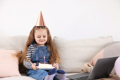 Photo of Birthday celebration. Little girl with piece of cake having video call via laptop on sofa at home
