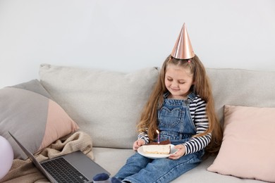 Photo of Birthday celebration. Little girl with piece of cake having video call via laptop on sofa at home