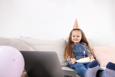 Photo of Birthday celebration. Little girl with piece of cake having video call via laptop on sofa at home