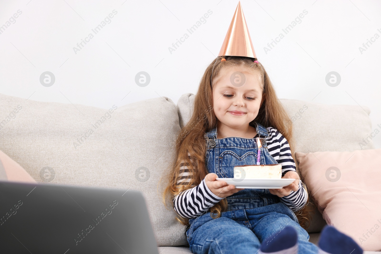 Birthday celebration. Little girl with piece of cake having video call via laptop on sofa at home Photo of Birthday celebration. Little girl with piece of cake having video call via laptop on sofa at home