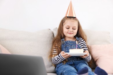 Photo of Birthday celebration. Little girl with piece of cake having video call via laptop on sofa at home