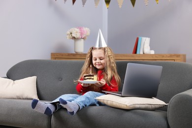 Photo of Birthday celebration. Little girl with piece of cake having video call via laptop on sofa at home
