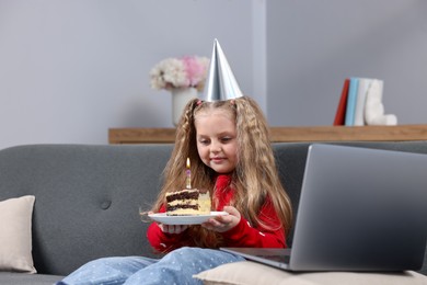 Photo of Birthday celebration. Little girl with piece of cake having video call via laptop on sofa at home
