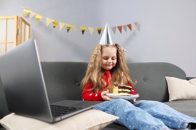 Photo of Birthday celebration. Little girl with piece of cake having video call via laptop on sofa at home