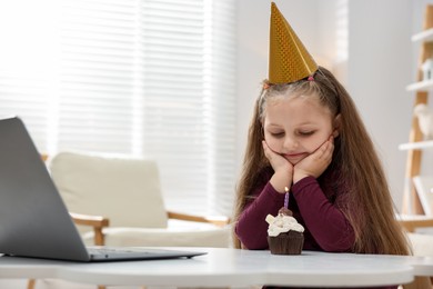 Photo of Birthday celebration. Little girl with cupcake having video call via laptop at table indoors