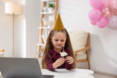 Photo of Birthday celebration. Little girl with cupcake having video call via laptop at table indoors