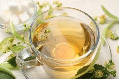 Photo of Fresh linden tea in cup, sugar cubes and flowers on white wooden table, closeup