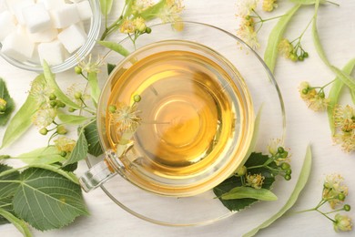 Photo of Fresh linden tea in cup, sugar cubes and flowers on white wooden table, flat lay