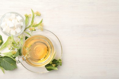 Photo of Fresh linden tea in cup, sugar cubes and flowers on white wooden table, flat lay. Space for text