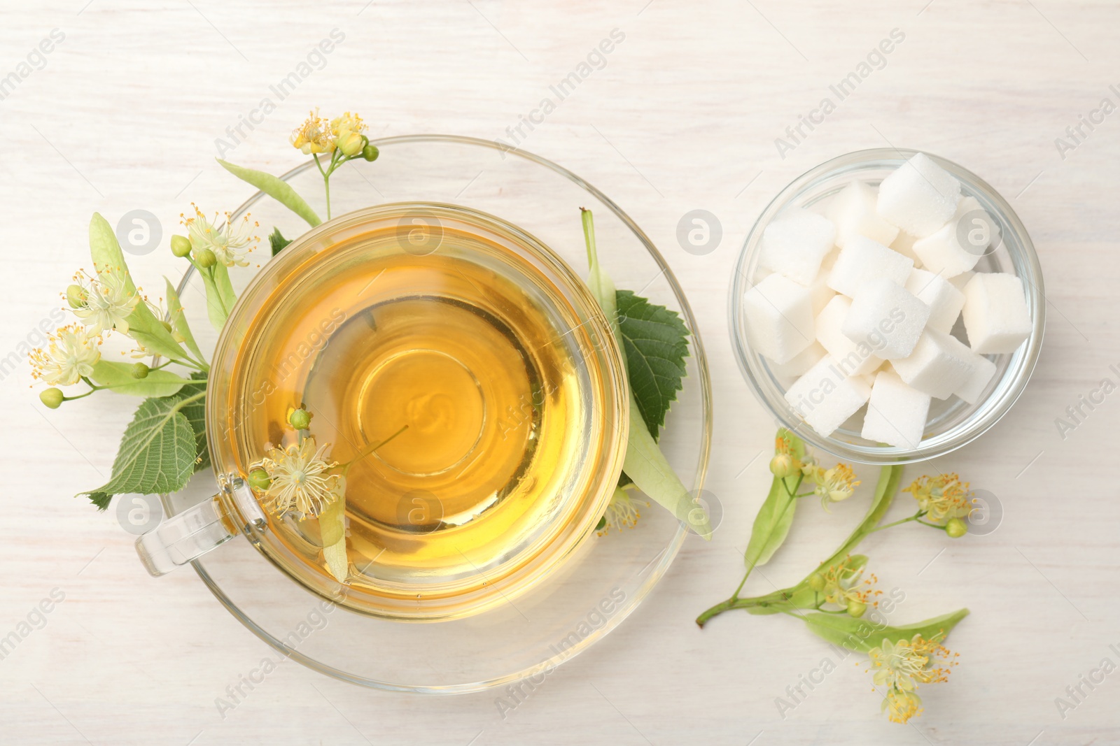 Photo of Fresh linden tea in cup, sugar cubes and flowers on white wooden table, flat lay